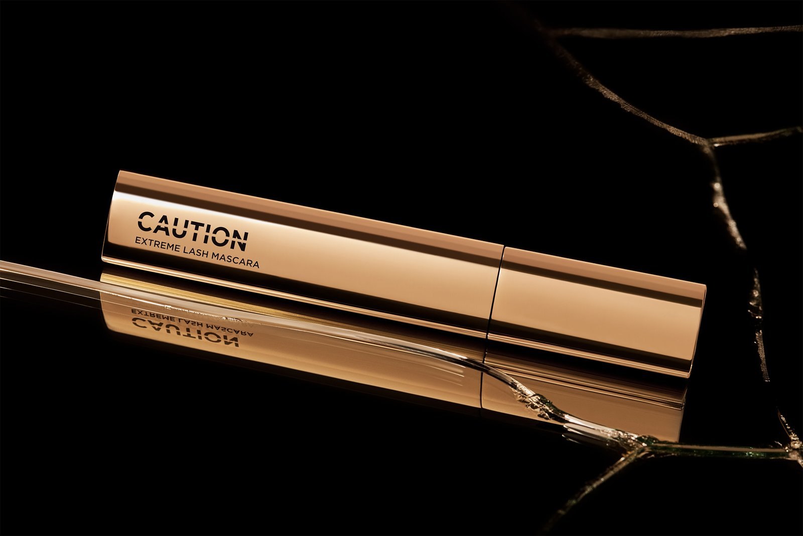 The gold CAUTION Extreme Lash Mascara on black with shards of broken glass